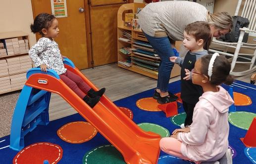 3yo at the top of a lil tykes sliding board is cheered on by both her big sister and another child at the workshop