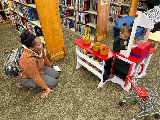 Adult caregiver watches while child plays at a market stand. 