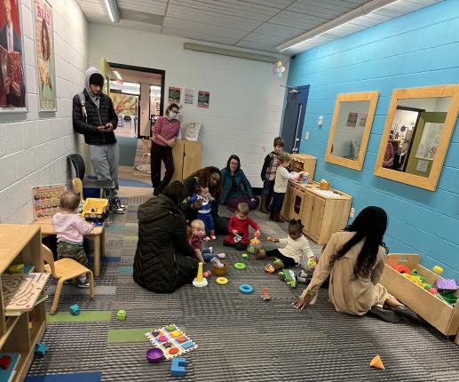 A photo of the Park Place Library KidZone where adults and babies are playing with toys on the floor and in the play kitchen area during our early literacy program.