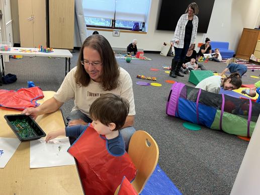 Parent and child finger painting together with other children and adults playing in the background.