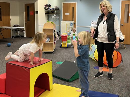 Caregiver looks on while a child pretends to take a photo of another child who is atop a climbing block. Other children playing in the toy kitchen in the background.