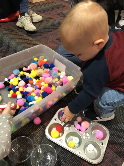 a toddler boy bending down to play with a bucket full of colorful pompoms