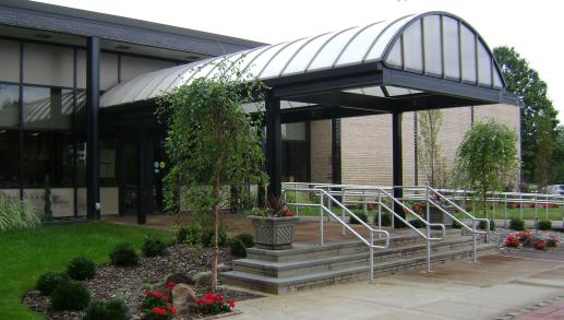 A view of the main entrance of the Henry Waldinger Memorial Library with steps, awning and surrounding plants and greenery in view.