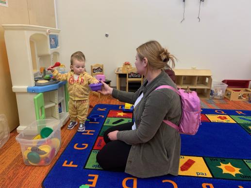 Mother is kneeling on the floor beside her 2 year old daughter playing cooking at a pretend kitchen set.  The little girl is holding a red spoon and handing her mom a purple bowl.