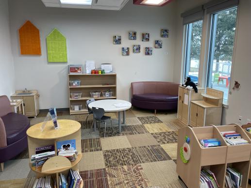 Library play area with two comfy benches, light wood kitchen set, shelves with toy bins, and board book bins. 