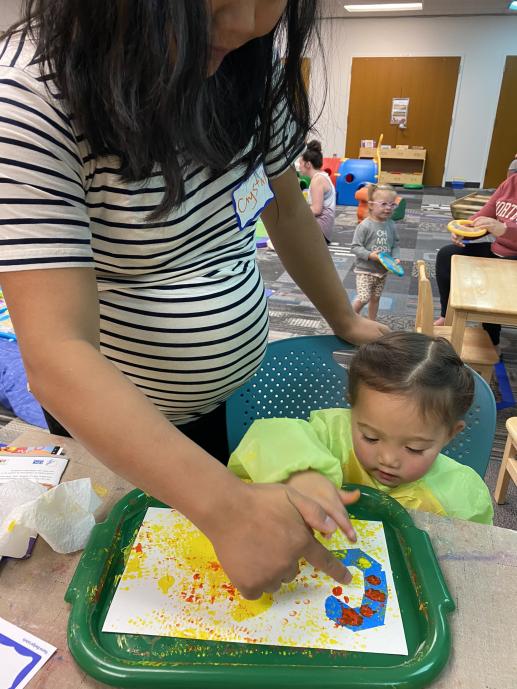 Mother and daughter at process art station, painting with corn cobs and pointing at a letter. In the background, others are playing with musical instruments.