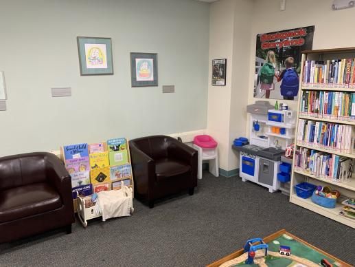 Two chairs accompany a small play space including a train table, play kitchen, high chair, and baby doll cradle. 