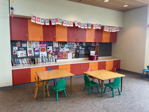 One wall of the children's section at the Stella Link library. There are small tables with little chairs for children to sit in. Kids artwork is displayed on a bulletin board behind the collection of parenting books. A banner reads "Every Child Is An Artist."