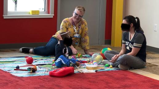 Librarian, mother and child sitting on carpet. Child is holding a musical instrument while the adults look on and smile. 