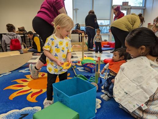 Little girl holding toy looking at mom holding baby on lap.  In background see several parents playing with children in various areas